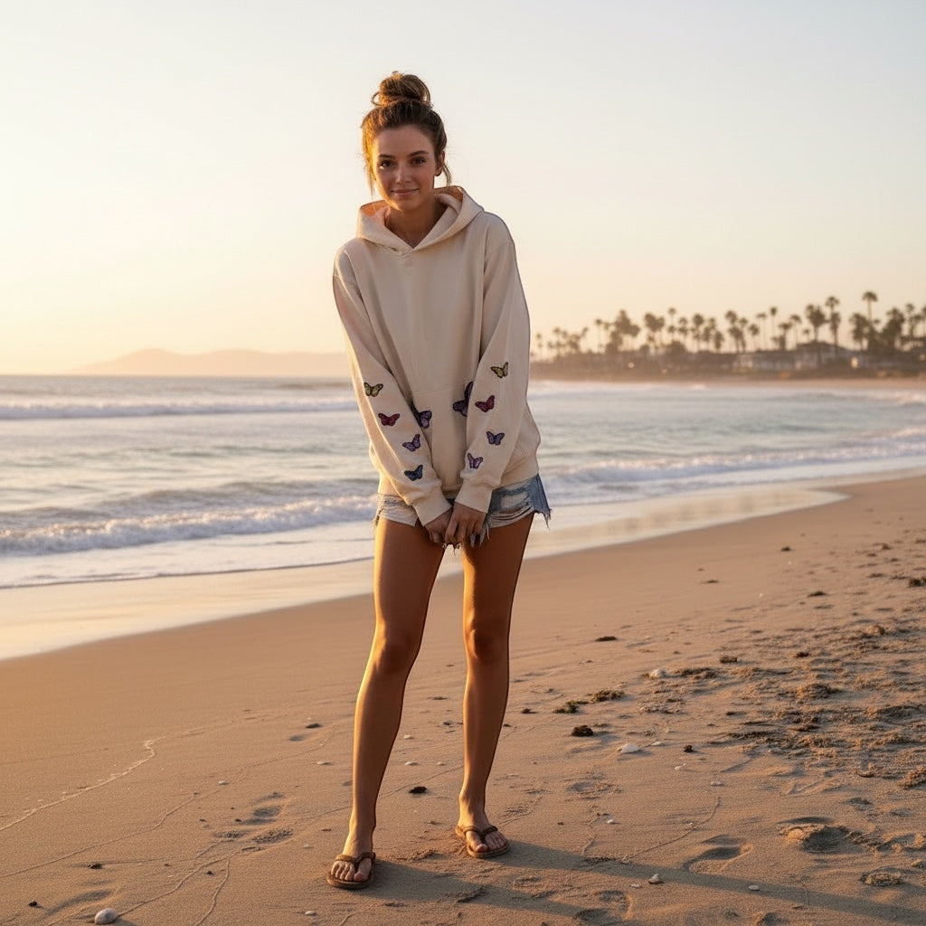 A Brand Called California cream unisex heavy cotton hoodie with embroidered butterflies on sleeves, worn by a woman on a beach at sunset.