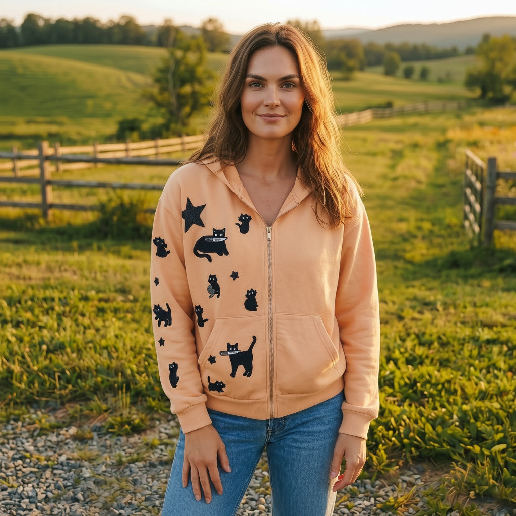 A Brand Called California peach cropped zip-up hoodie with black cat and star appliques. Model wears jeans in a field.